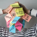 Una persona agobiada por notas adhesivas con tareas: "Kids", "Bills", "Homework", "Work", "Shopping", "Gym", "School", "Groceries", "Cleaning". Un recordatorio visual de cómo ser mejor persona es priorizar.