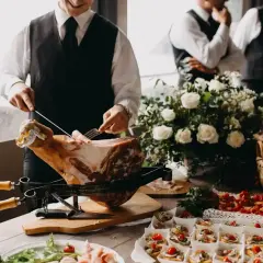 Un hombre corta jamón para montar un catering en casa. Hay canapés, flores y otros invitados en el fondo.