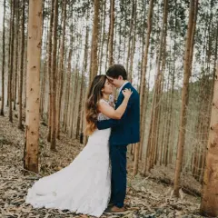 Pareja abrazándose en un bosque de eucaliptos. Ella lleva un vestido de novia blanco, él un traje azul. Una escena romántica, quizás un "trash the dress" post-boda.
