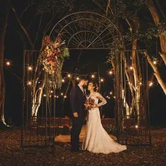 Pareja en un arco adornado con flores, bajo luces de feria. ¿Es una boda, no boda?