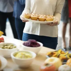 Una mujer lleva una bandeja de pastelitos para una merienda de cumpleaños para adultos. Hay langosta, tomates y otros aperitivos en la mesa.