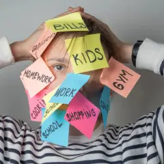 Una persona agobiada por notas adhesivas con tareas: "Kids", "Bills", "Homework", "Work", "Shopping", "Gym", "School", "Groceries", "Cleaning". Un recordatorio visual de cómo ser mejor persona es priorizar.