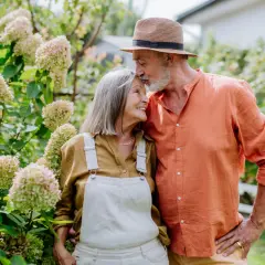 Pareja mayor en un jardín, él le da un beso en la frente. Sus gestos expresan los lenguajes del amor.