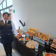 Una mujer sonriente supervisa un montaje de coffee break con frutas, postres, pan y agua.