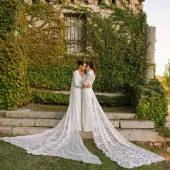 Dos novias se abrazan frente a una pared de piedra cubierta de hiedra, luciendo impresionantes ajuares de novia con velos largos y encaje.