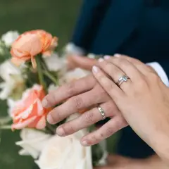 Manos entrelazadas con anillos, celebrando aniversarios de boda junto a un ramo de flores color melocotón y blanco.