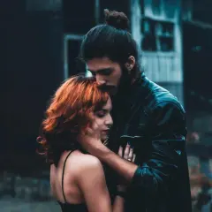 Un hombre besa la frente de una mujer con cabello rojizo. La escena evoca la complejidad de la infidelidad emocional, un vínculo secreto que florece en la sombra.