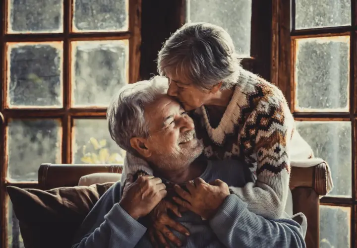 Una pareja madura se abraza tiernamente frente a una ventana. Ella le da un beso en la frente, él sonríe con ternura.