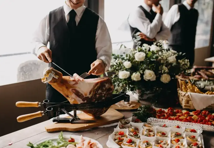 Un hombre corta jamón para montar un catering en casa. Hay canapés, flores y otros invitados en el fondo.