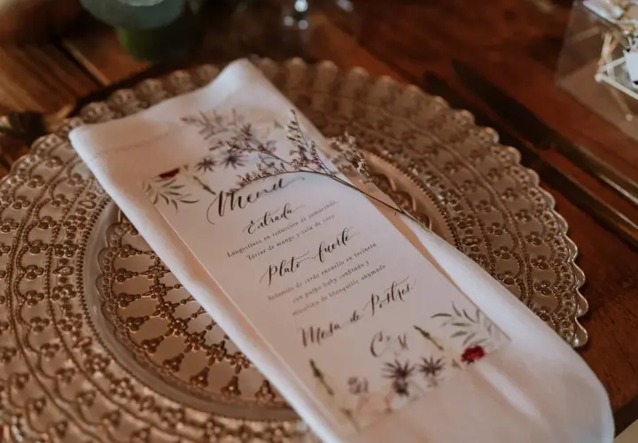Un elegante menú de boda con caligrafía floral detalla los platos: langostinos, solomito de cerdo y mesa de postres.