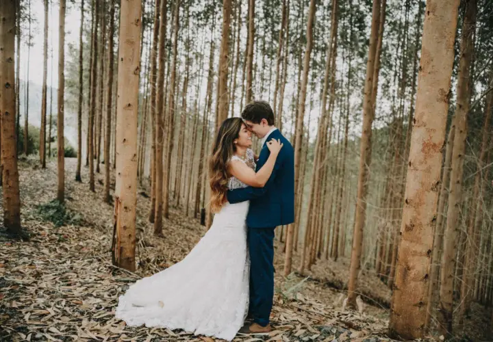 Pareja abrazándose en un bosque de eucaliptos. Ella lleva un vestido de novia blanco, él un traje azul. Una escena romántica, quizás un "trash the dress" post-boda.