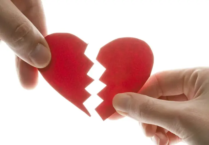 Manos separando un corazón rojo roto, símbolo del mal de amores.