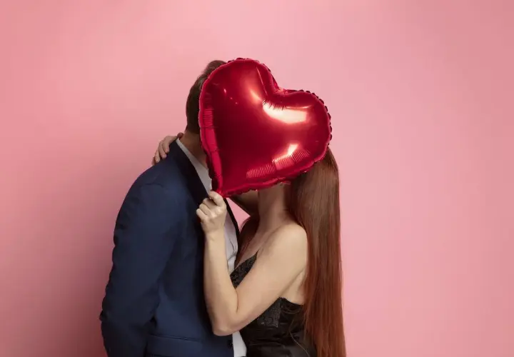 Pareja besándose detrás de un globo rojo en forma de corazón. Ideas románticas para sorprender a tu pareja.