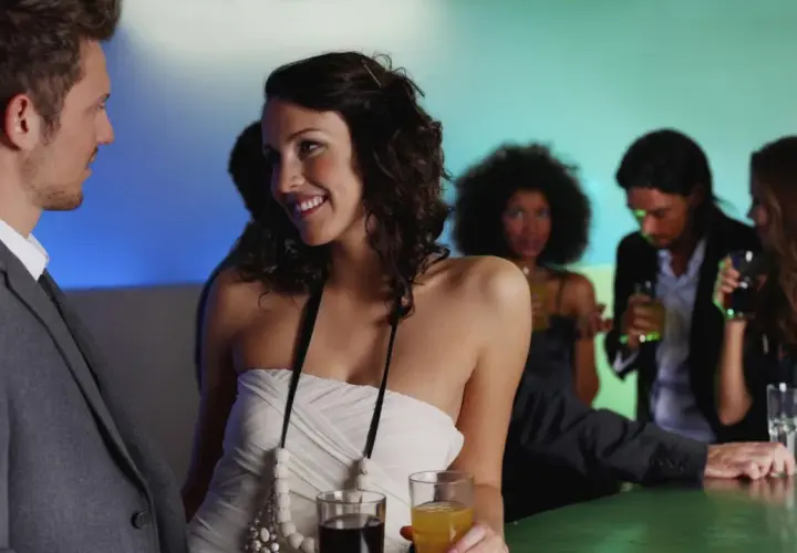 Pareja joven sonriendo y disfrutando de su primera cita en un bar, con otras personas de fondo.
