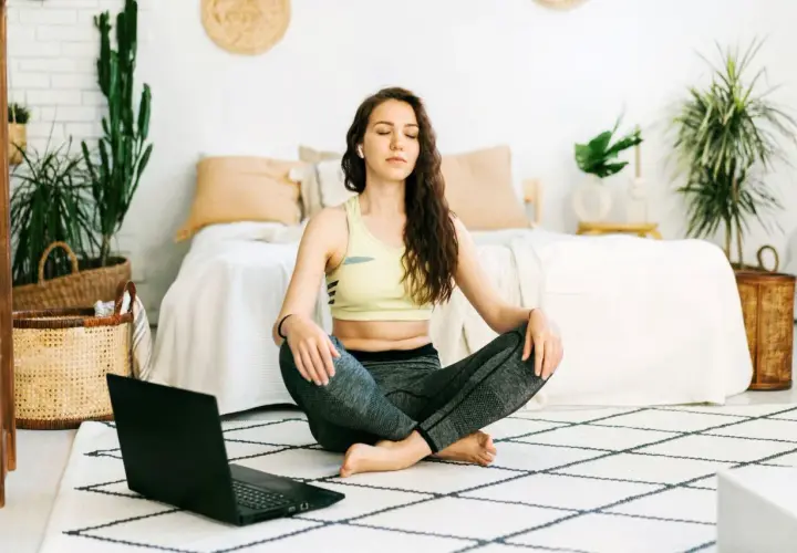 Mujer meditando en posición de loto, aprendiendo técnicas de relajación con un portátil.
