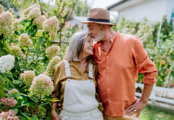 Pareja mayor en un jardín, él le da un beso en la frente. Sus gestos expresan los lenguajes del amor.