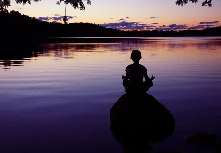 Silueta de persona meditando en una roca al atardecer, buscando la paz interior junto a un lago sereno.