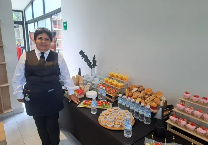 Una mujer sonriente supervisa un montaje de coffee break con frutas, postres, pan y agua.