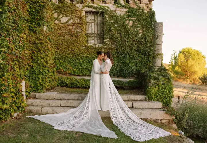 Dos novias se abrazan frente a una pared de piedra cubierta de hiedra, luciendo impresionantes ajuares de novia con velos largos y encaje.