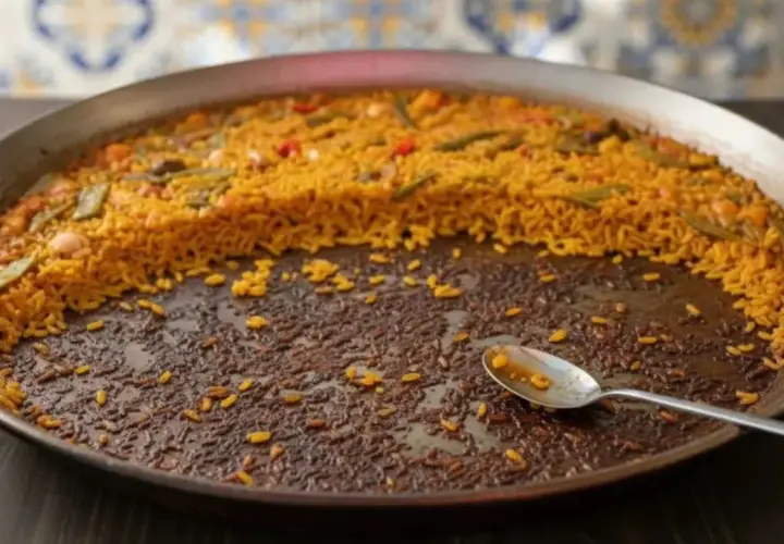 Paella casi terminada en una paellera. El arroz pegado en el fondo, con una cuchara, indica que ha sido deliciosa.