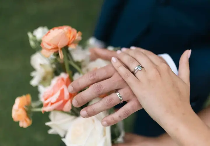 Manos entrelazadas con anillos, celebrando aniversarios de boda junto a un ramo de flores color melocotón y blanco.