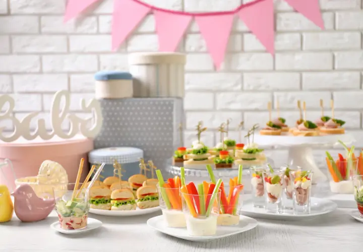 Mesa de baby shower con deliciosa comida: mini hamburguesas, canapés, palitos de verduras con dip y vasitos de aperitivos.