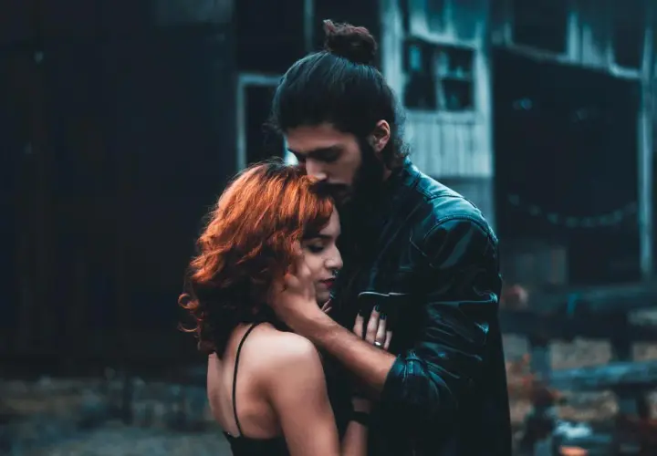 Un hombre besa la frente de una mujer con cabello rojizo. La escena evoca la complejidad de la infidelidad emocional, un vínculo secreto que florece en la sombra.