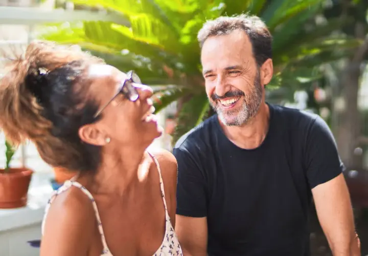 Pareja riendo al aire libre. Él, superando la crisis de los 50, sonríe con complicidad. Ella, con gafas de sol, se ríe a carcajadas.
