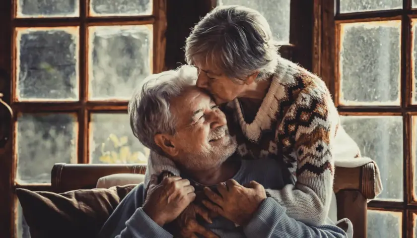 Una pareja madura se abraza tiernamente frente a una ventana. Ella le da un beso en la frente, él sonríe con ternura.