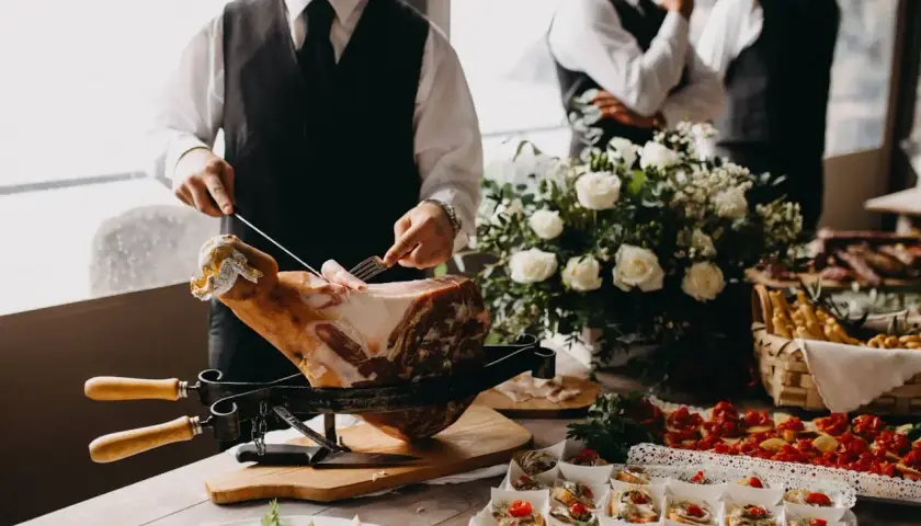 Un hombre corta jamón para montar un catering en casa. Hay canapés, flores y otros invitados en el fondo.