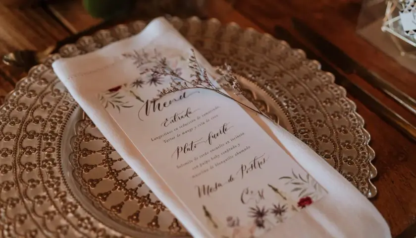Un elegante menú de boda con caligrafía floral detalla los platos: langostinos, solomito de cerdo y mesa de postres.