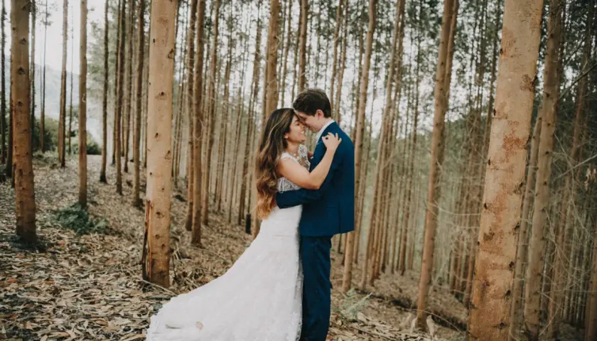 Pareja abrazándose en un bosque de eucaliptos. Ella lleva un vestido de novia blanco, él un traje azul. Una escena romántica, quizás un "trash the dress" post-boda.