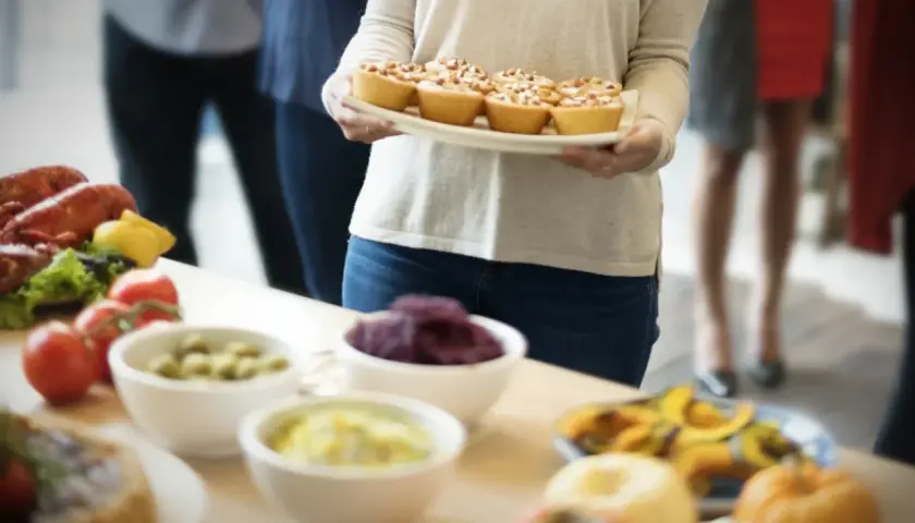 Una mujer lleva una bandeja de pastelitos para una merienda de cumpleaños para adultos. Hay langosta, tomates y otros aperitivos en la mesa.