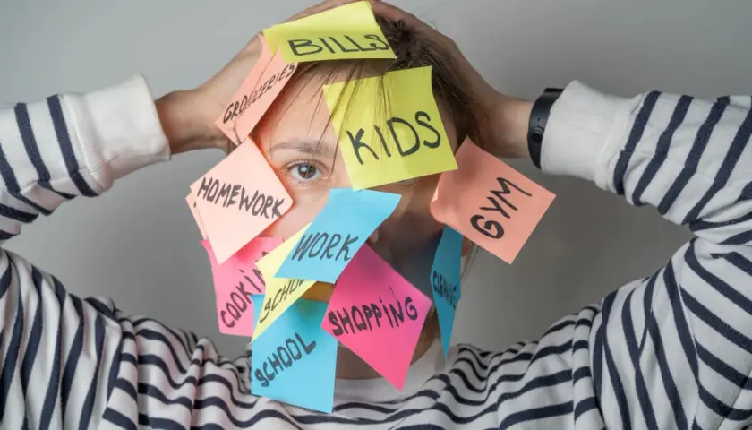 Una persona agobiada por notas adhesivas con tareas: "Kids", "Bills", "Homework", "Work", "Shopping", "Gym", "School", "Groceries", "Cleaning". Un recordatorio visual de cómo ser mejor persona es priorizar.