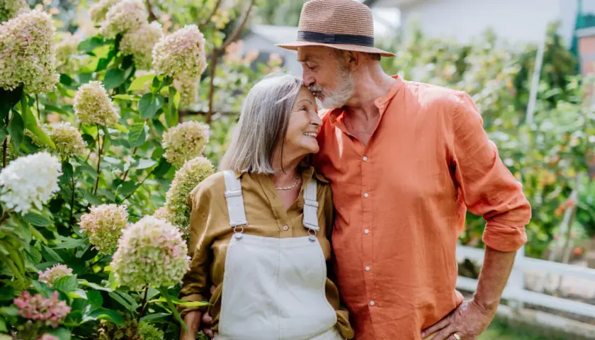 Pareja mayor en un jardín, él le da un beso en la frente. Sus gestos expresan los lenguajes del amor.