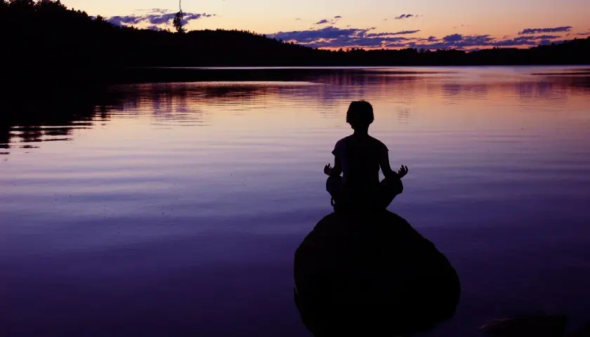 Silueta de persona meditando en una roca al atardecer, buscando la paz interior junto a un lago sereno.