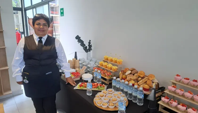 Una mujer sonriente supervisa un montaje de coffee break con frutas, postres, pan y agua.