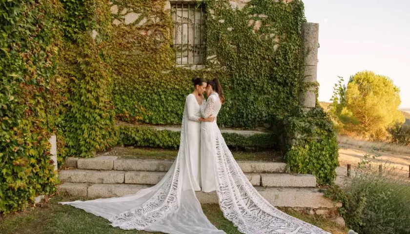 Dos novias se abrazan frente a una pared de piedra cubierta de hiedra, luciendo impresionantes ajuares de novia con velos largos y encaje.