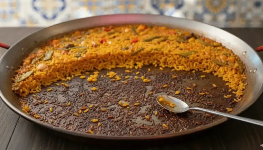 Paella casi terminada en una paellera. El arroz pegado en el fondo, con una cuchara, indica que ha sido deliciosa.