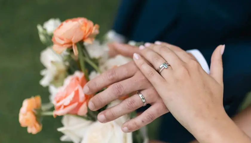 Manos entrelazadas con anillos, celebrando aniversarios de boda junto a un ramo de flores color melocotón y blanco.
