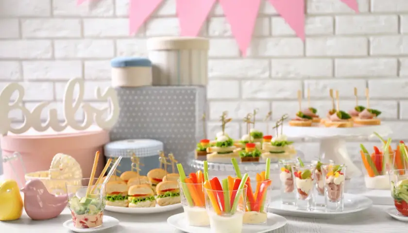 Mesa de baby shower con deliciosa comida: mini hamburguesas, canapés, palitos de verduras con dip y vasitos de aperitivos.