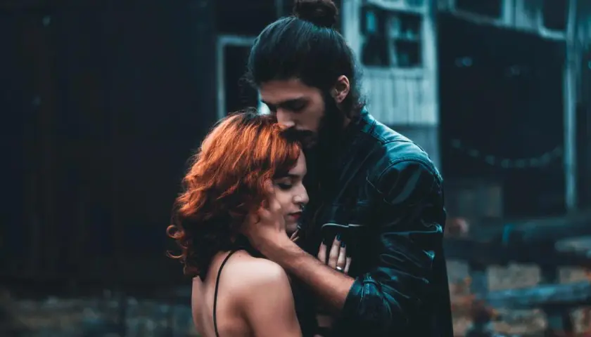Un hombre besa la frente de una mujer con cabello rojizo. La escena evoca la complejidad de la infidelidad emocional, un vínculo secreto que florece en la sombra.