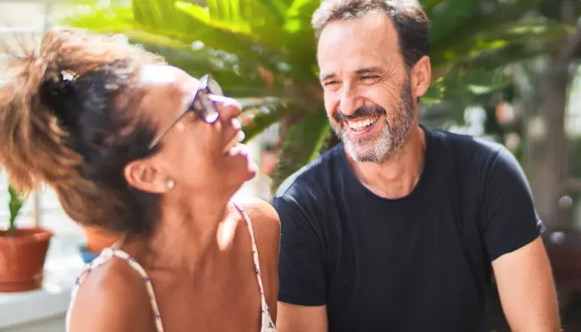 Pareja riendo al aire libre. Él, superando la crisis de los 50, sonríe con complicidad. Ella, con gafas de sol, se ríe a carcajadas.