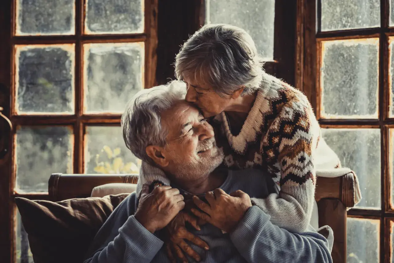 Una pareja madura se abraza tiernamente frente a una ventana. Ella le da un beso en la frente, él sonríe con ternura.
