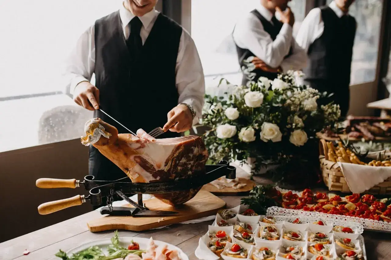Un hombre corta jamón para montar un catering en casa. Hay canapés, flores y otros invitados en el fondo.