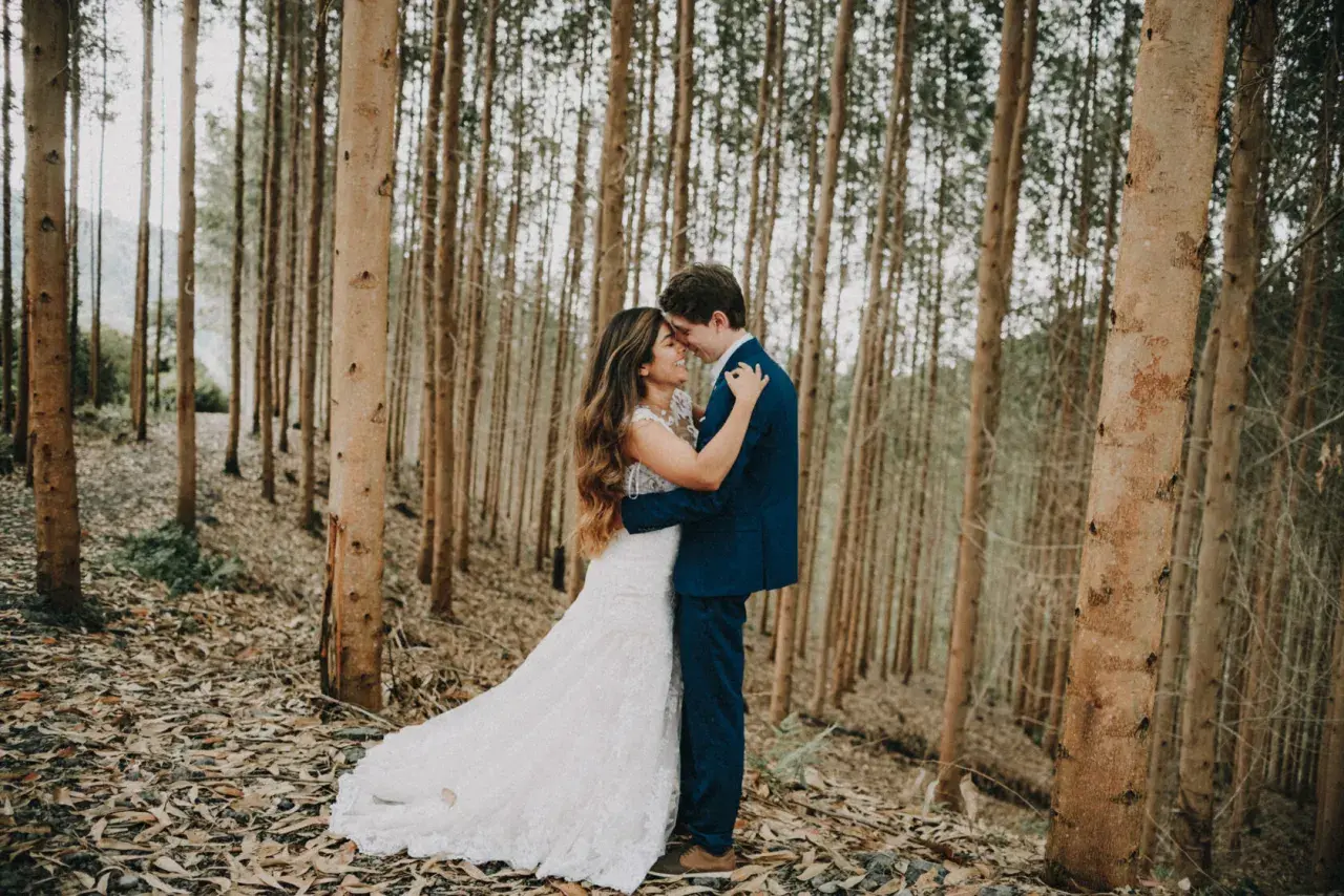 Pareja abrazándose en un bosque de eucaliptos. Ella lleva un vestido de novia blanco, él un traje azul. Una escena romántica, quizás un "trash the dress" post-boda.