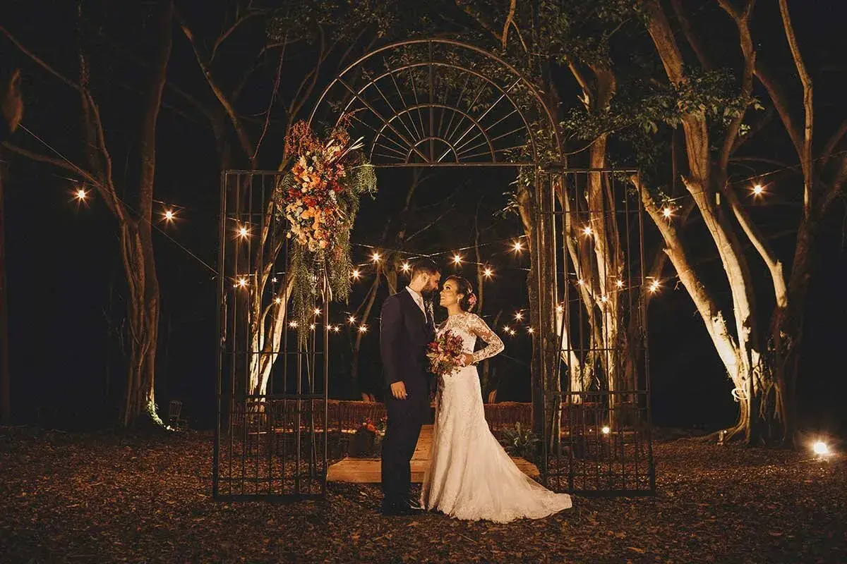 Pareja en un arco adornado con flores, bajo luces de feria. ¿Es una boda, no boda?