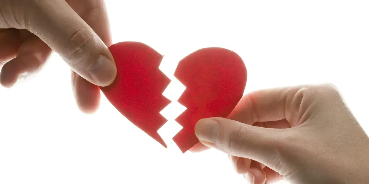 Manos separando un corazón rojo roto, símbolo del mal de amores.