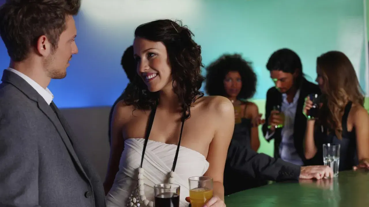 Pareja joven sonriendo y disfrutando de su primera cita en un bar, con otras personas de fondo.