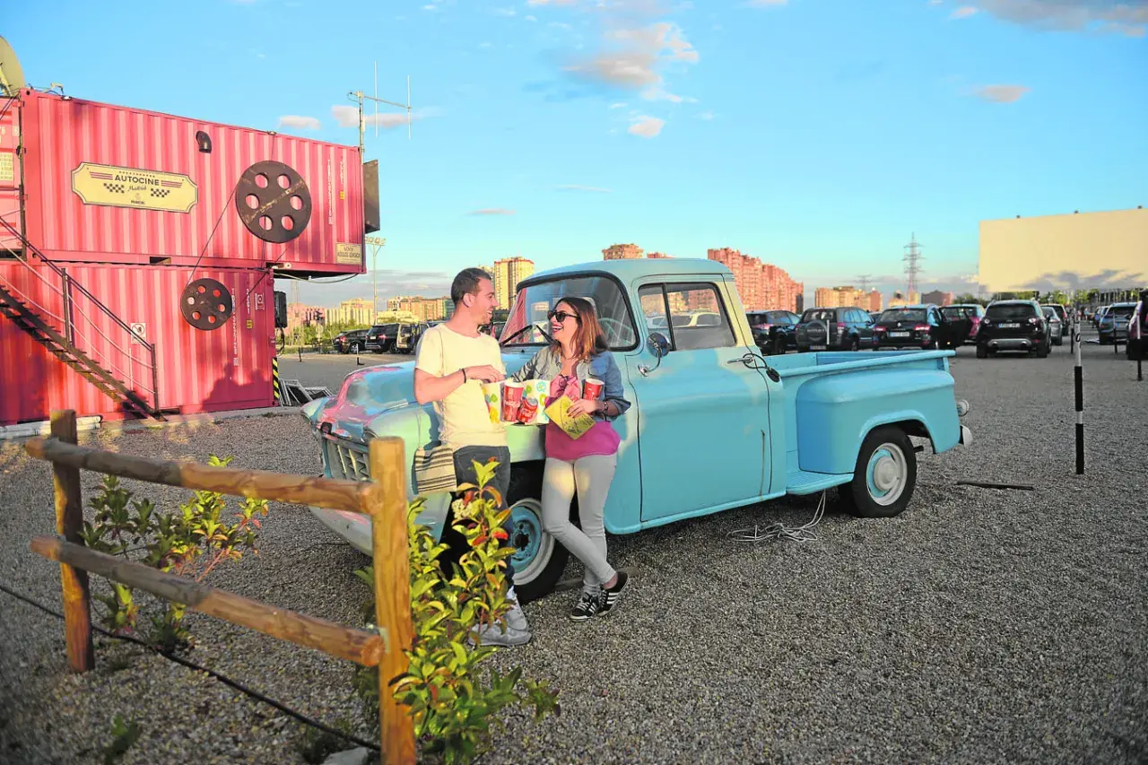 Pareja disfrutando de planes en pareja originales sin dinero en un autocine, apoyados en una camioneta azul, con palomitas y bebidas.